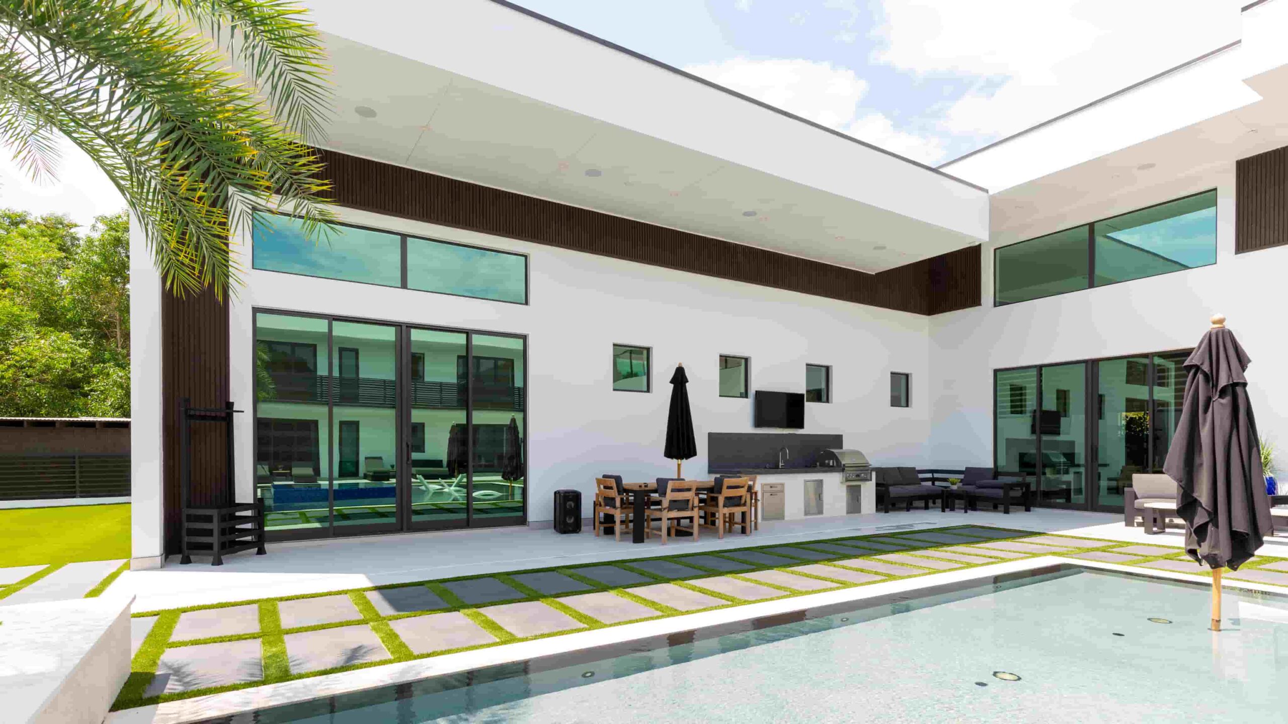 Modern house patio with a pool, outdoor dining area, lounge chairs, and large windows. Grass and stone walkway surrounds the pool. Two black umbrellas provide shade.