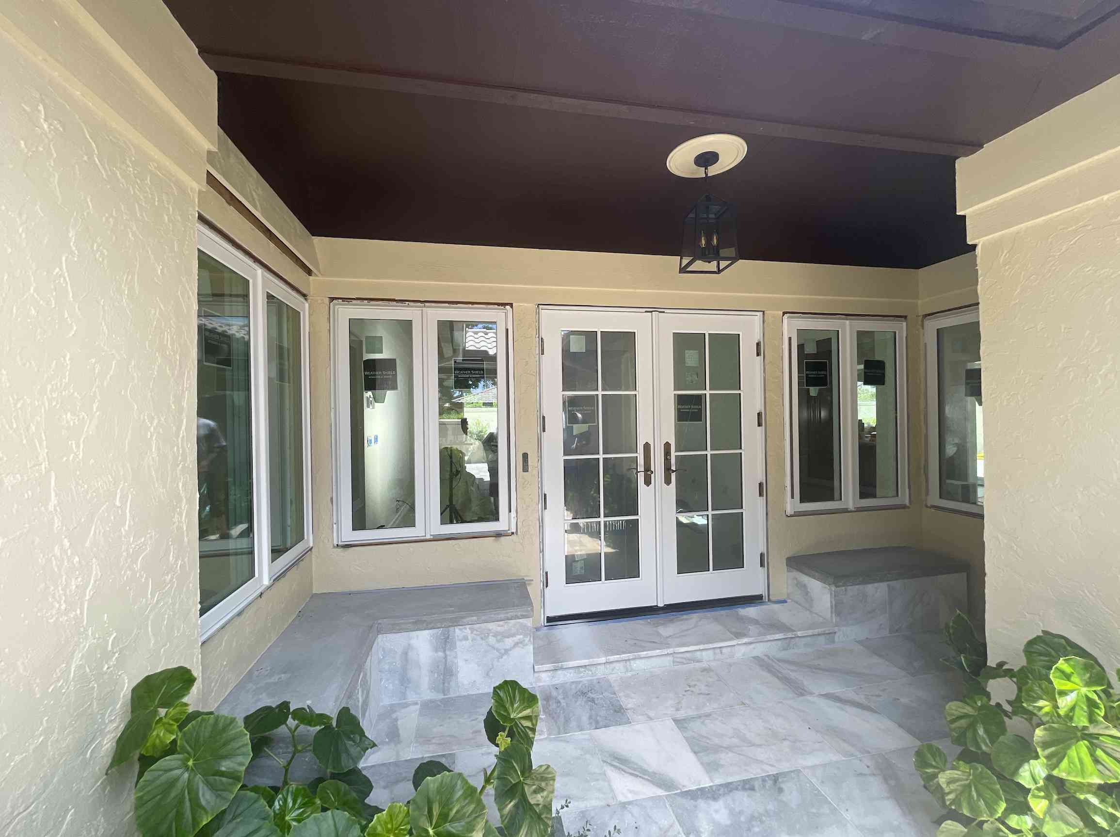 A beige house entryway with white double doors, sidelights, and multiple windows, flanked by green plants and featuring a lantern-style ceiling light.