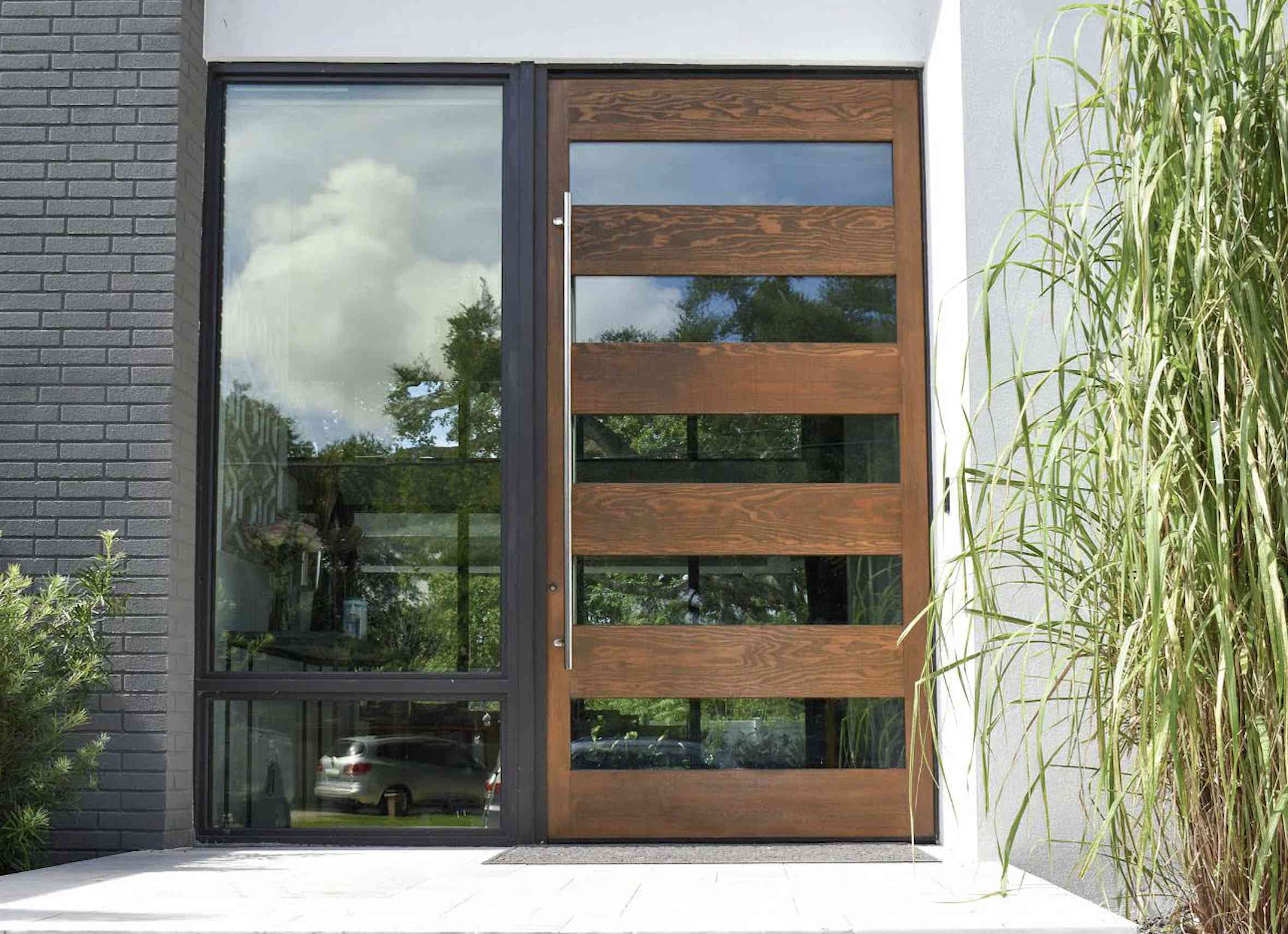 Modern hurricane door with horizontal wooden panels next to a large glass window, reflecting trees and sky; tall grass grows on the right side of the entrance.