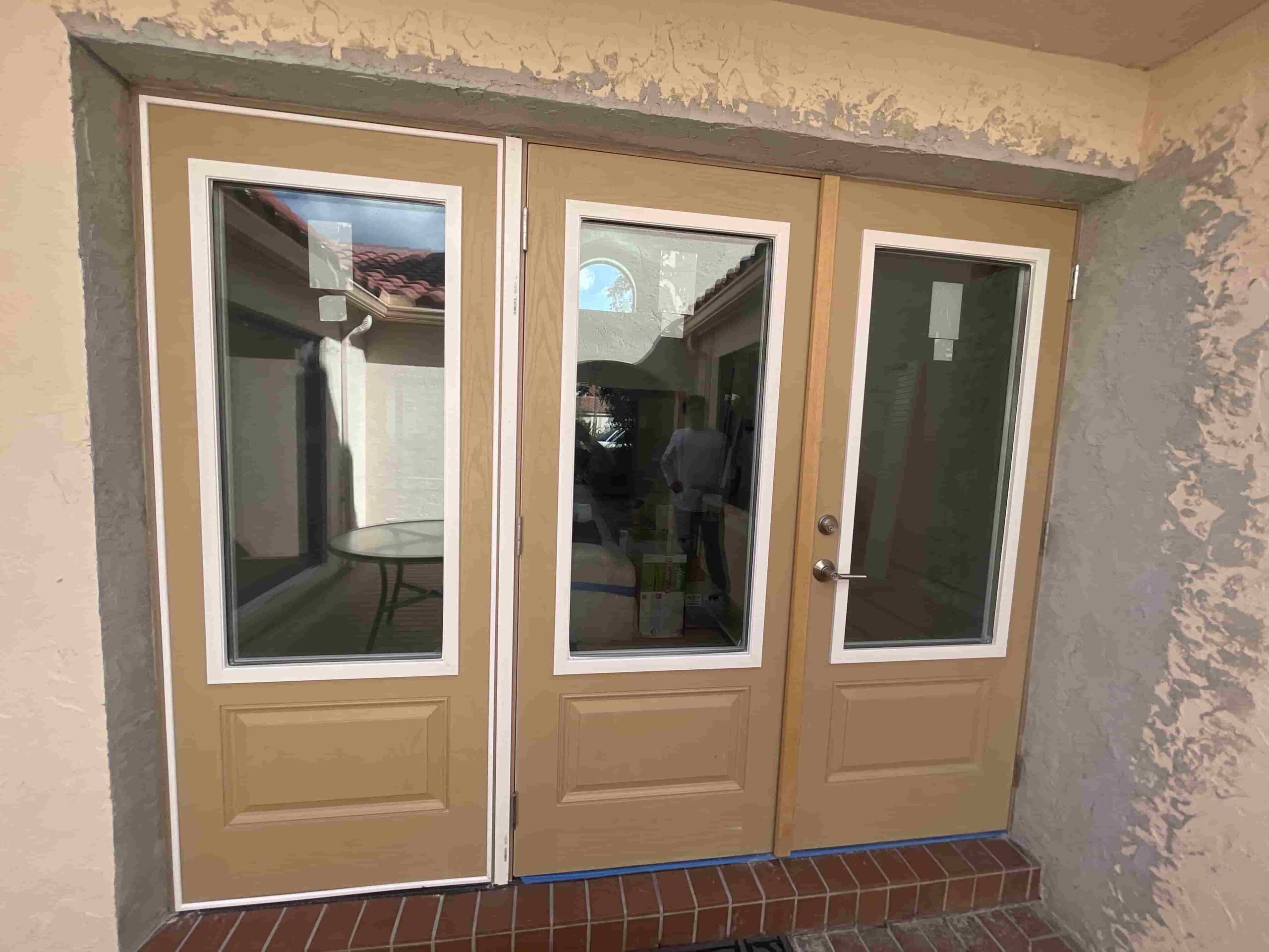 A set of tan double doors with glass panels, flanked by a matching narrow window section, opening onto a brick patio area outside a house.