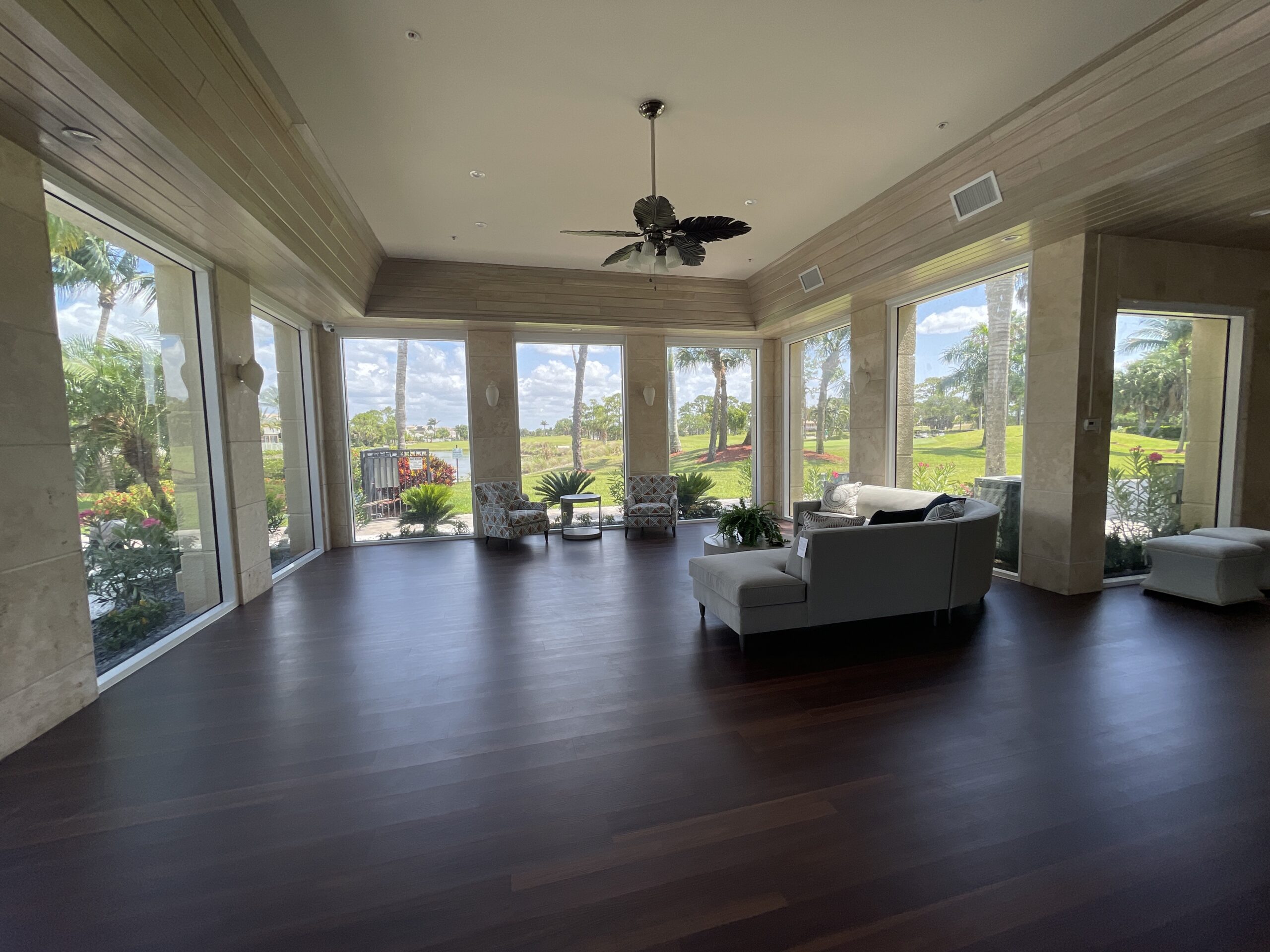 Spacious sunroom with large floor-to-ceiling windows, dark wood flooring, white furniture, potted plants, and views of a garden and trees outside.
