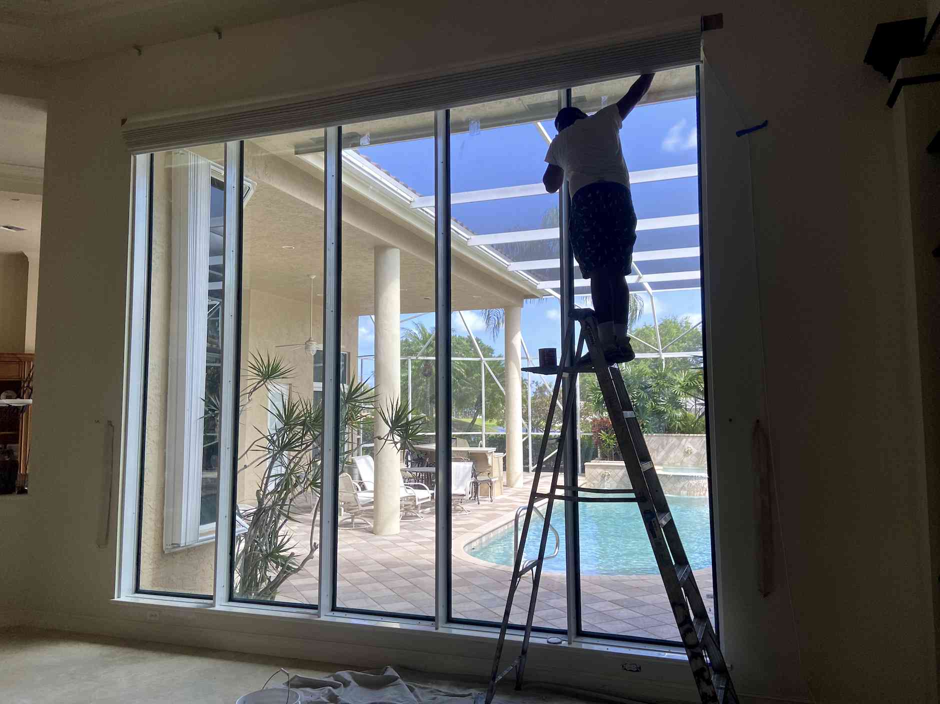 A person stands on a ladder indoors, working near a hurricane window overlooking a patio with a pool and plants outside.