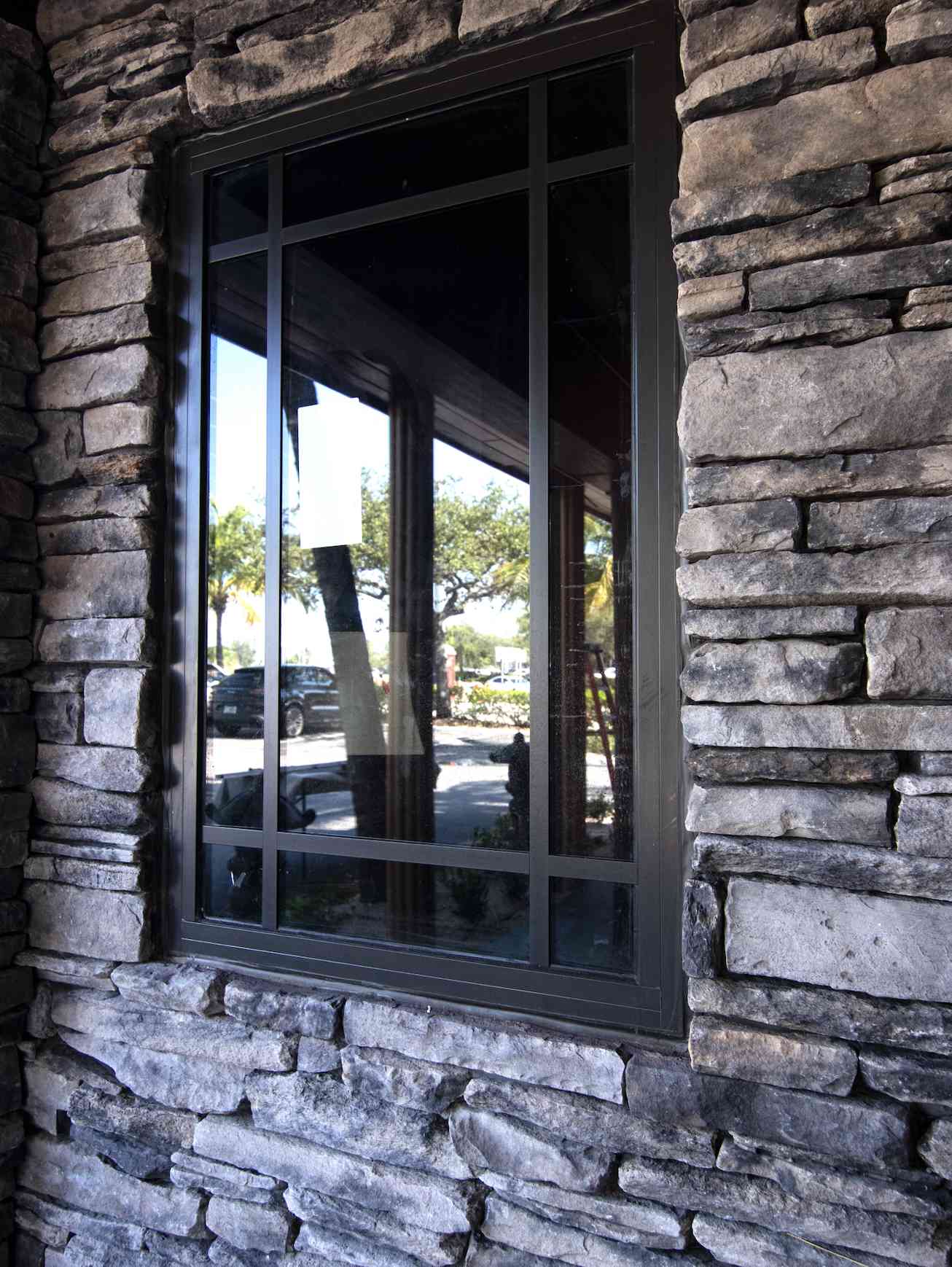 A rectangular window with a black frame is set in a gray stacked stone wall, reflecting trees and parked cars outside.