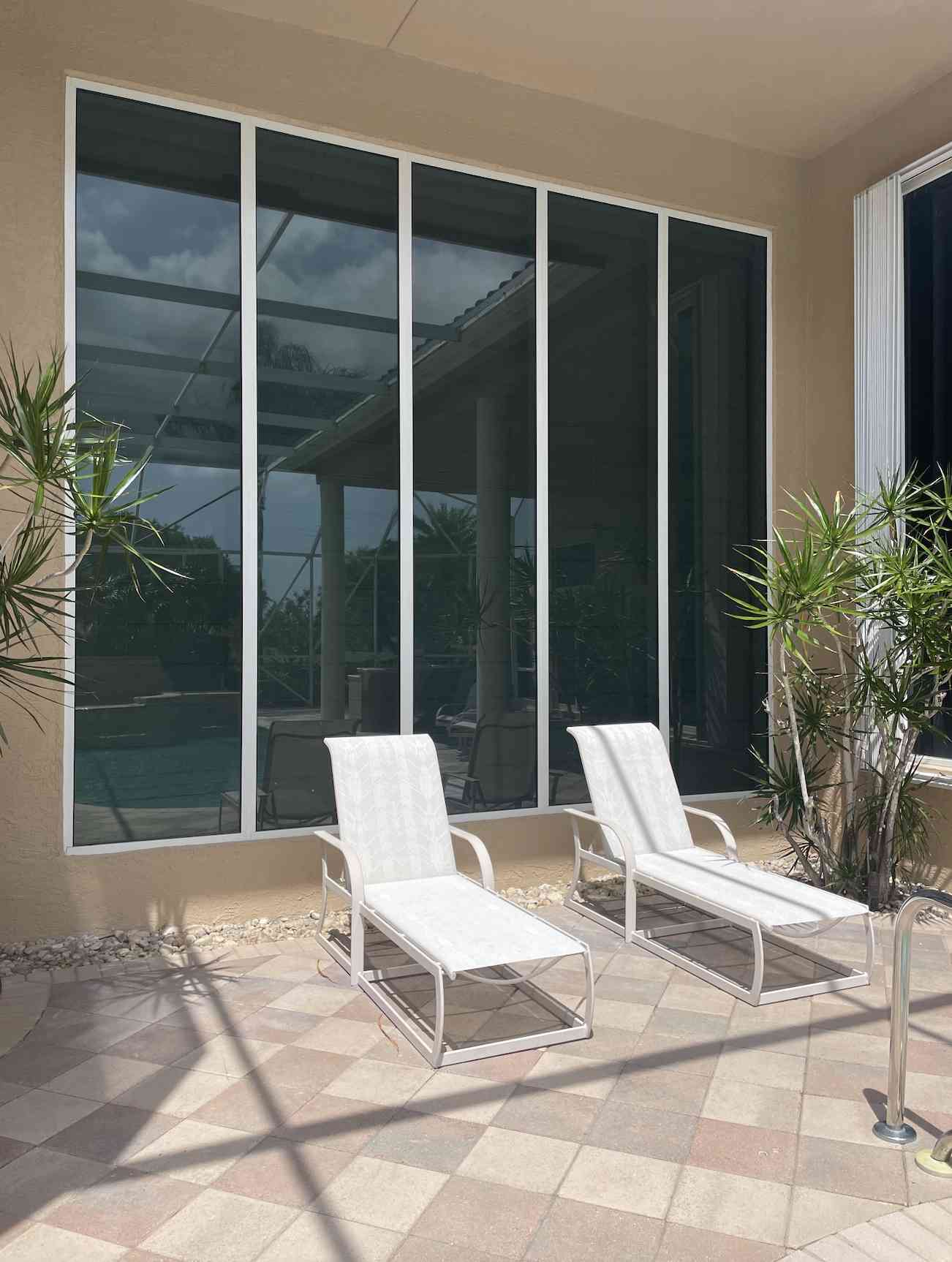 Two white lounge chairs are positioned on a tiled patio beside a large window, with potted plants nearby and sunlight casting shadows.