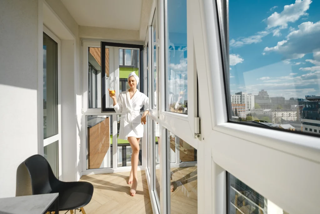 A person in a white bathrobe and towel stands on a balcony holding a drink, with large windows and a cityscape visible in the background on a sunny day.