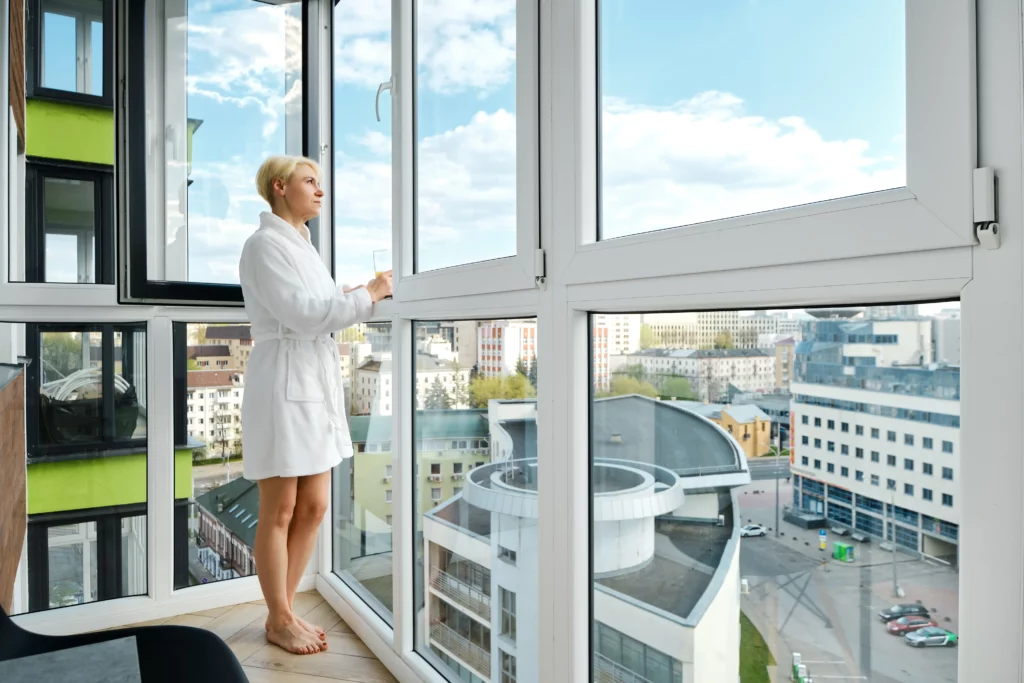 A woman in a white bathrobe stands on a balcony and looks out at an urban cityscape through large glass windows on a sunny day.