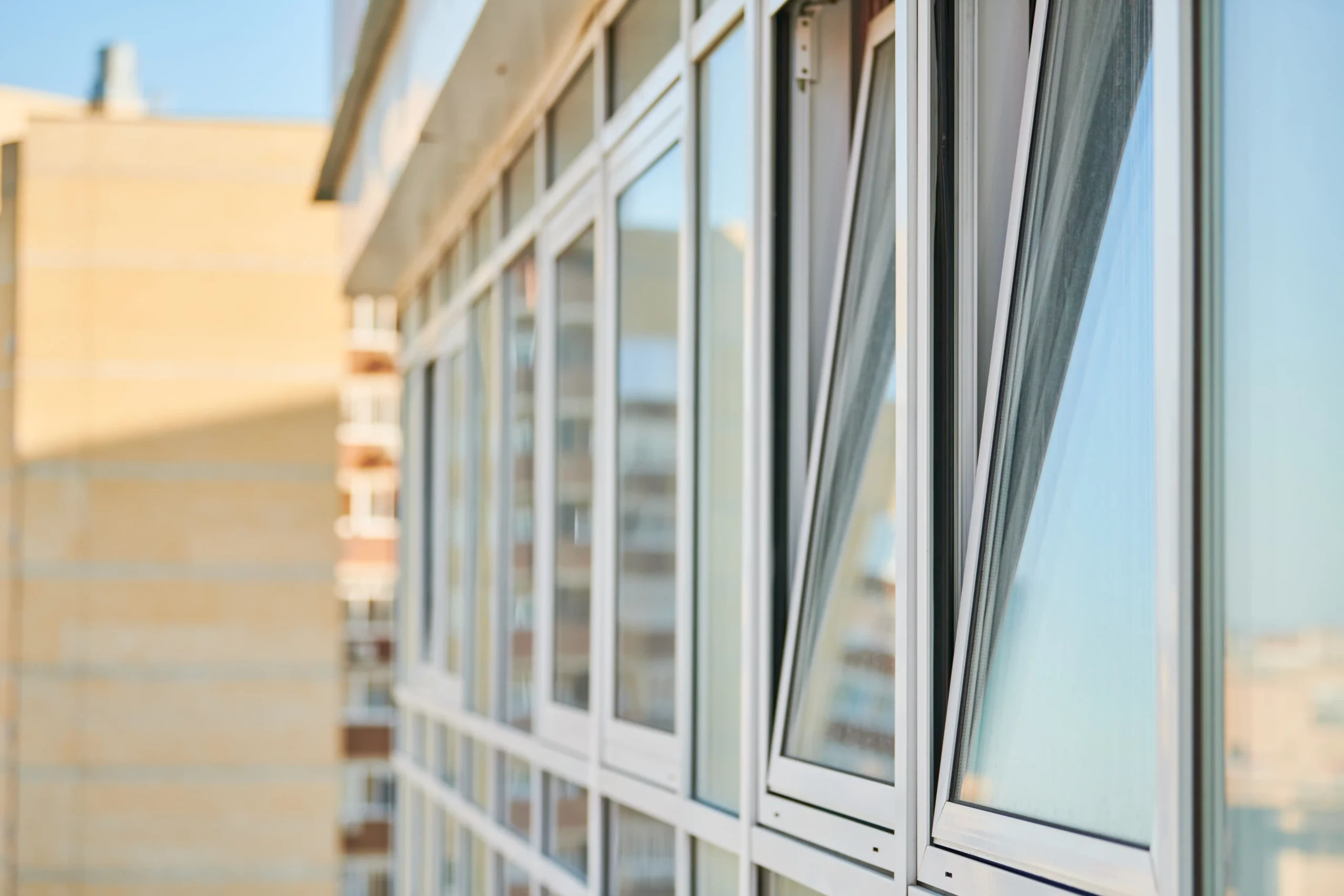 A close-up view of modern building windows, with several windows tilted open and blurred city buildings visible in the background.