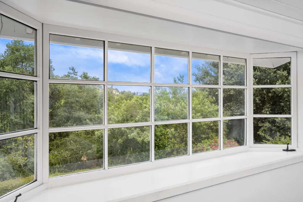 Large white-framed bay window overlooking green trees and shrubs under a blue sky with scattered clouds.